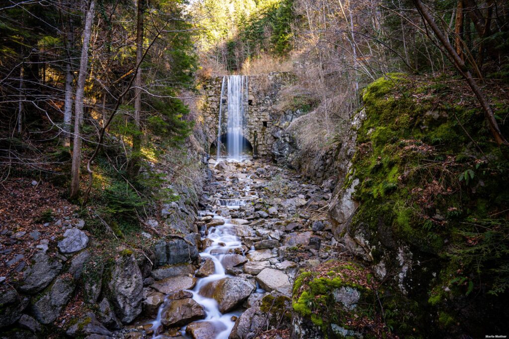 Ein Wasserfall stürzt am Gruonbach in Flüelen, im Kanton Uri, herab. Der Wasserfall ist von einer üppigen Vegetation umgeben, mit grünen Bäumen und Farnen, die entlang des Bachlaufs wachsen.