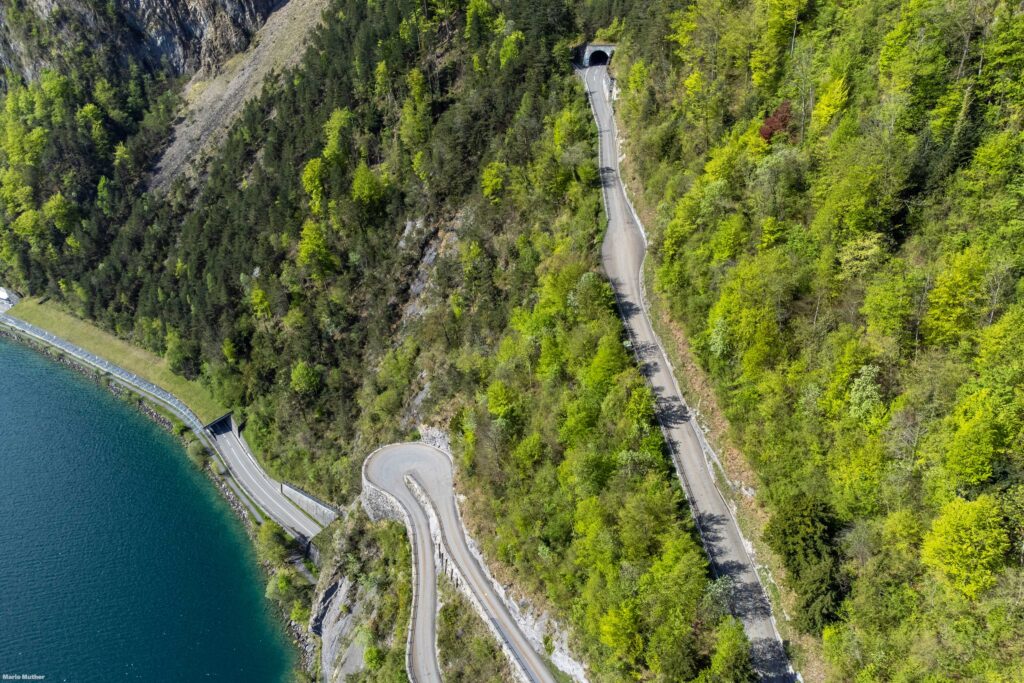 Die Drohnenfotografie zeigt die Isenthaler-Bergstrasse im Kanton Uri. Die Strasse windet sich steil den Hang hinauf und bietet einen atemberaubenden Blick auf die umliegenden Berge und Täler.